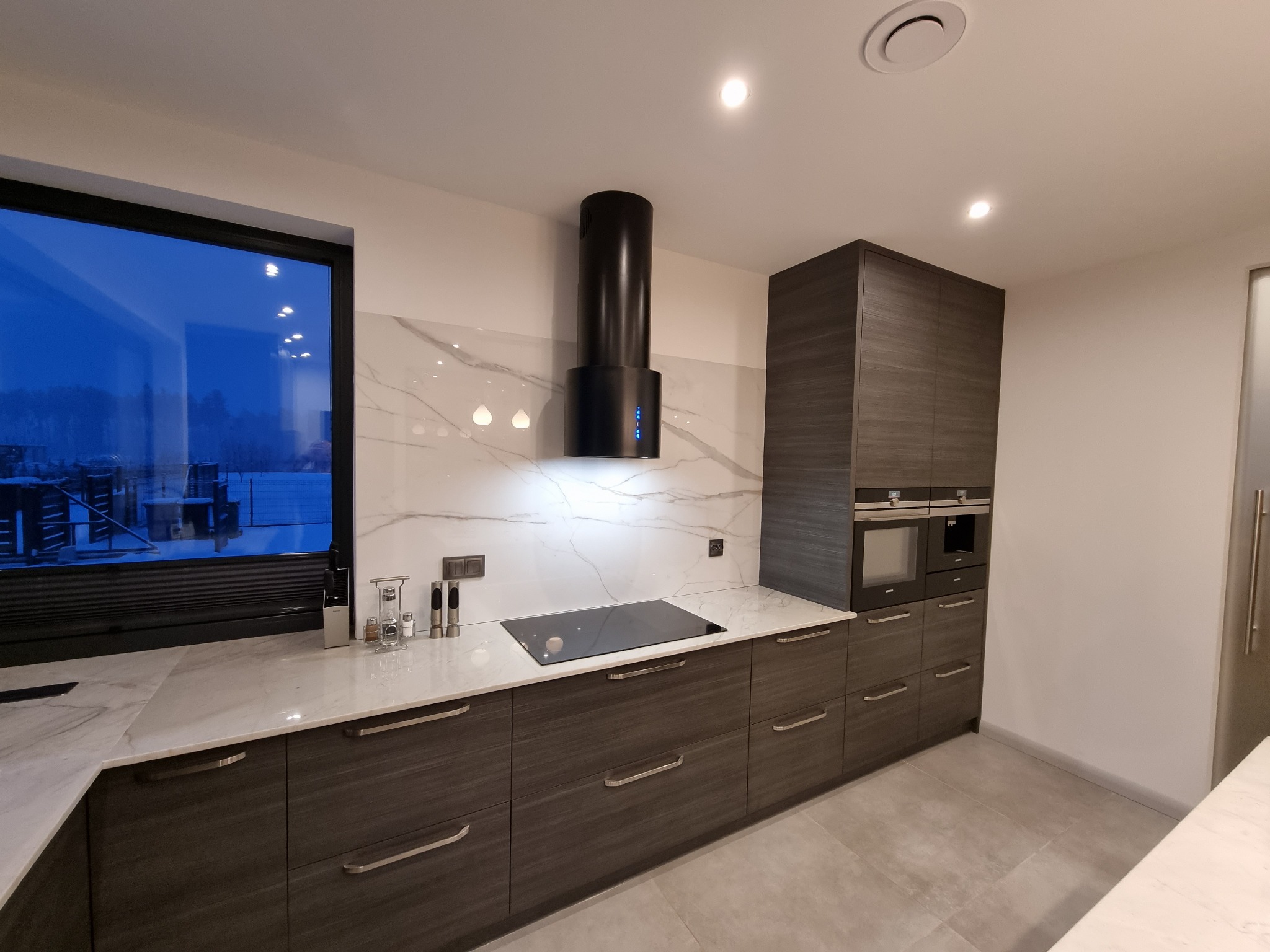 Nowoczesna kuchnia z ciemnymi, drewnopodobnymi frontami szafek, białym blatem z marmurowym wzorem i czarnym okapem cylindrycznym, widok na zimowy krajobraz za oknem.