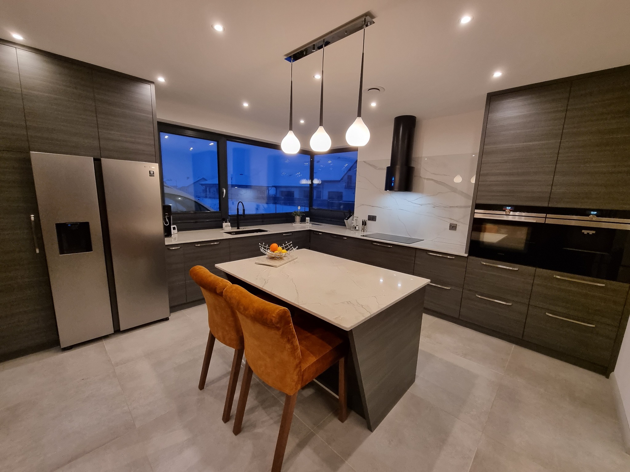 Nowoczesna kuchnia z ciemnymi, drewnopodobnymi frontami, wyspą z marmurowym blatem i pomarańczowymi krzesłami barowymi pod trzema wiszącymi lampami.
