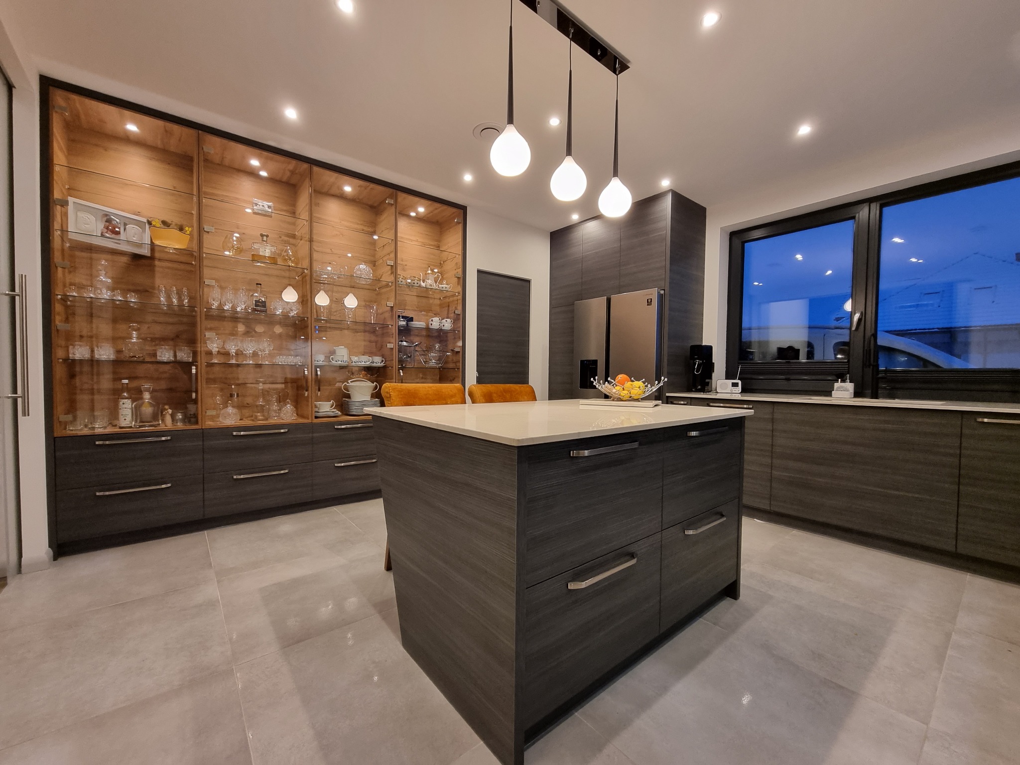Nowoczesna kuchnia z wyspą, ciemnoszare szafki z drewnianym wzorem, przeszklona witryna z oświetleniem i dekoracyjnymi przedmiotami, trzy lampy wiszące nad wyspą, duże okno z widokiem na zmierzch.