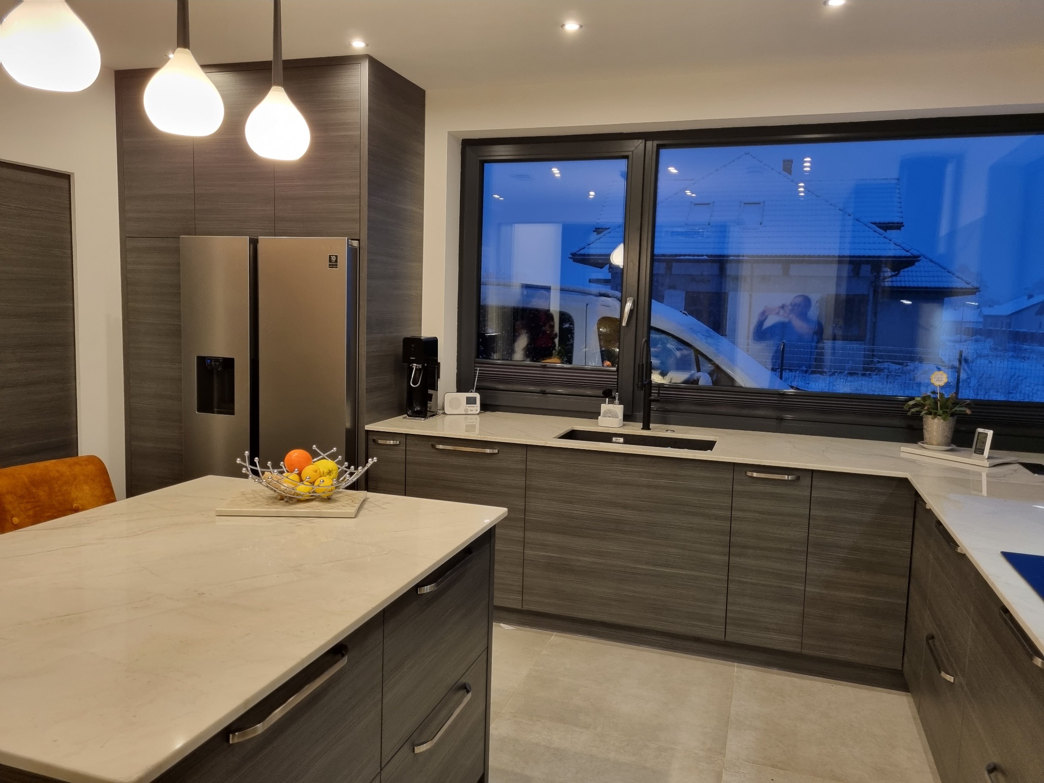 Nowoczesna kuchnia z szarymi szafkami z drewnianą teksturą, białym blatem, wyspą kuchenną z owocami, dużym oknem z widokiem na zasypany śniegiem dom i osobą robiącą zdjęcie, oświetlona trzema...