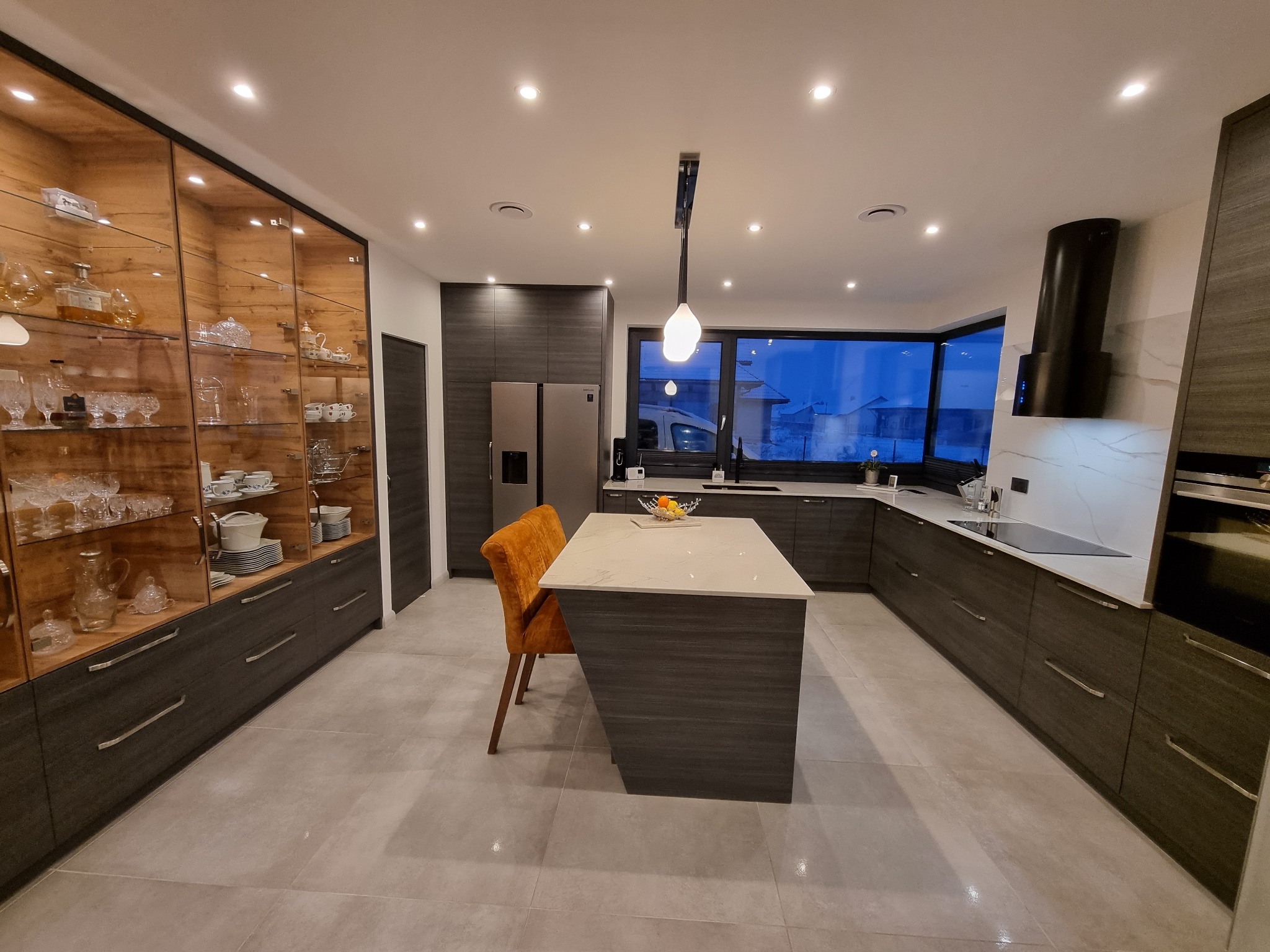 Nowoczesna kuchnia z ciemnymi, drewnopodobnymi szafkami, wyspą z blatem imitującym marmur, i przeszkloną witryną z zastawą stołową, oświetlona punktowymi lampami sufitowymi.