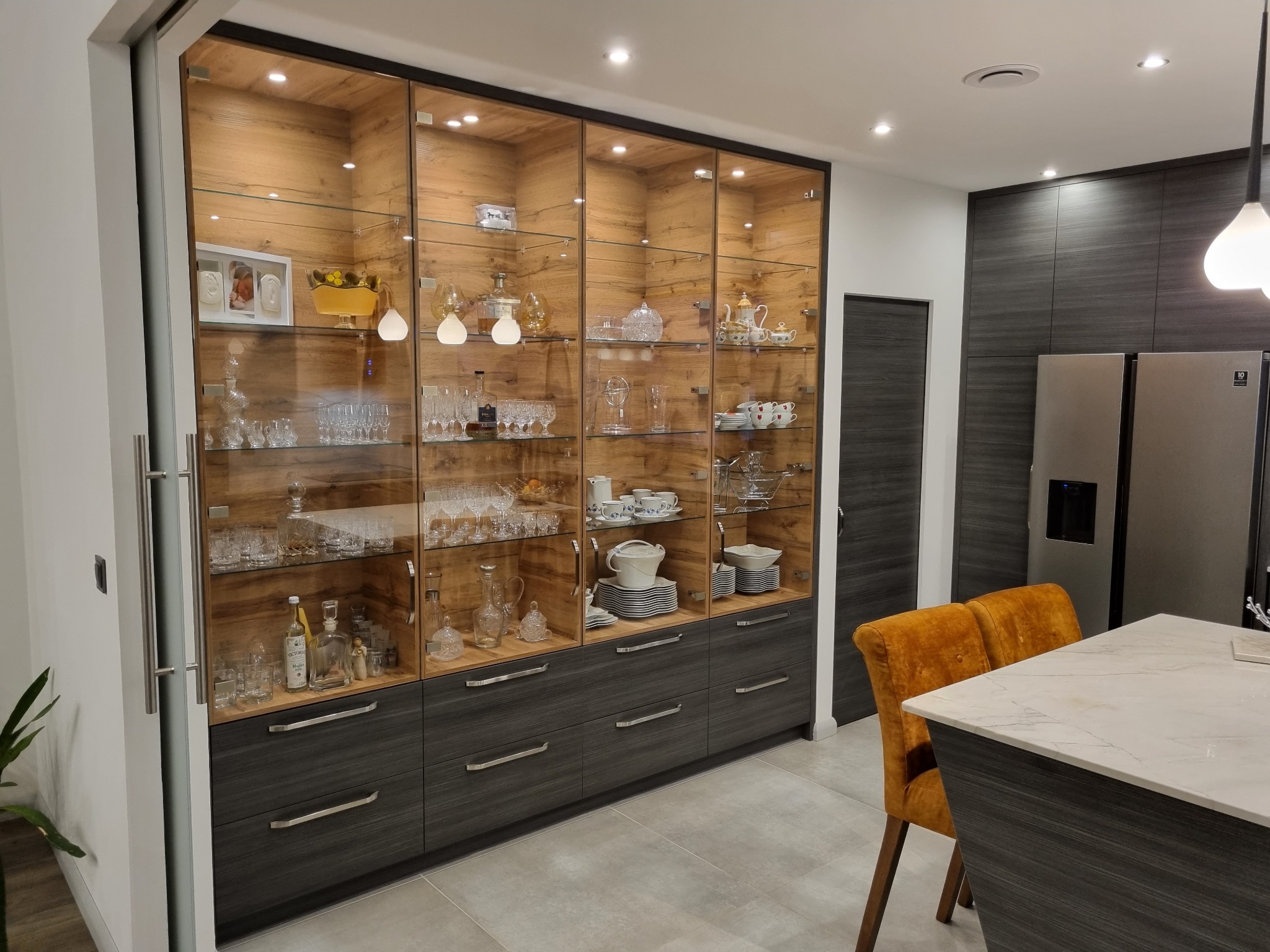 Wysoka przeszklona szafa w zabudowie z szufladami w dolnej części, podświetlona, wypełniona szkłem, porcelaną i dekoracjami, w nowoczesnej kuchni z szarymi frontami i wyspą.