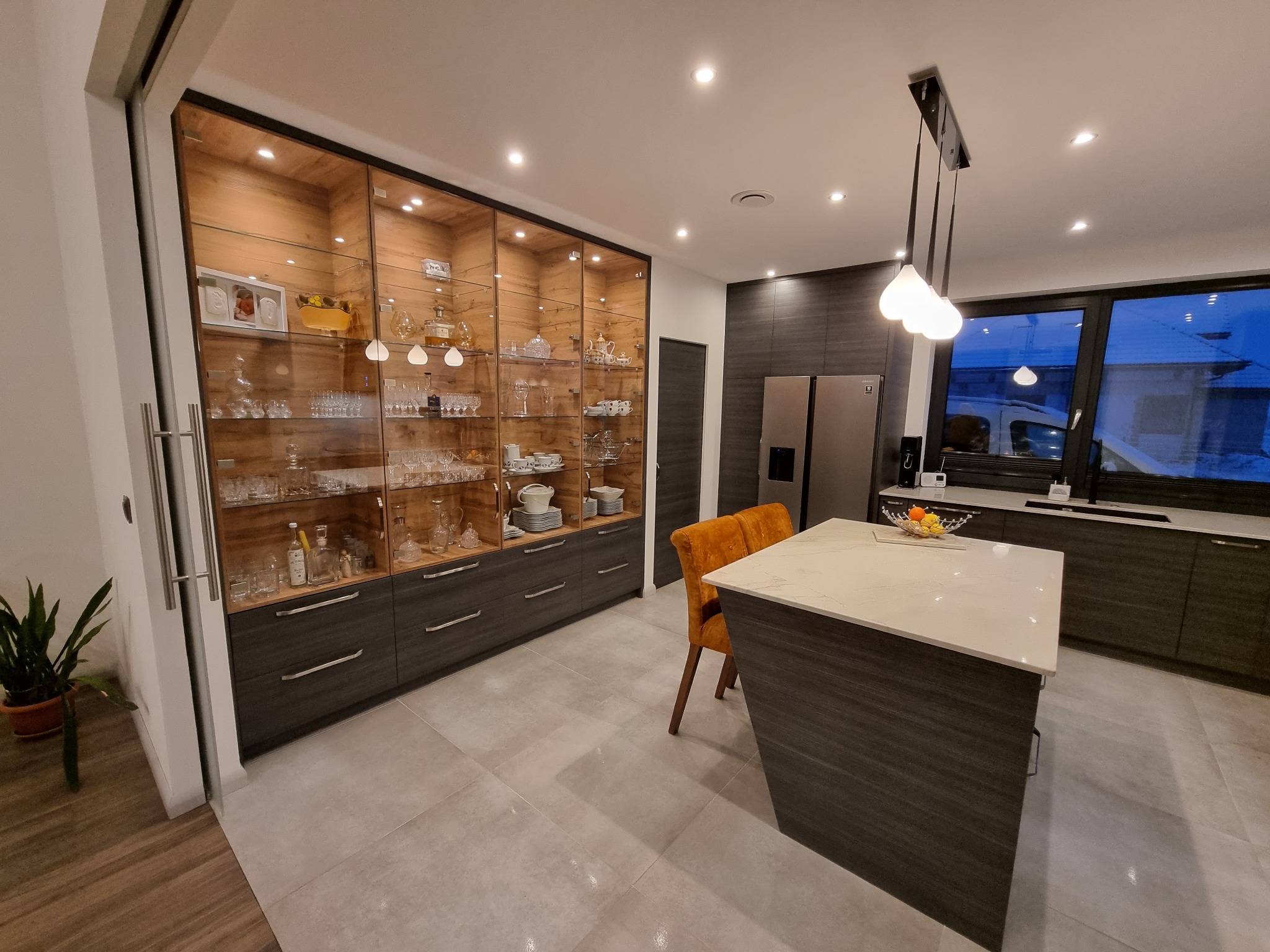 Nowoczesna kuchnia z szarymi szafkami, wyspą z białym blatem i szklaną witryną z drewnianym wnętrzem, eksponującą kryształy i porcelanę.