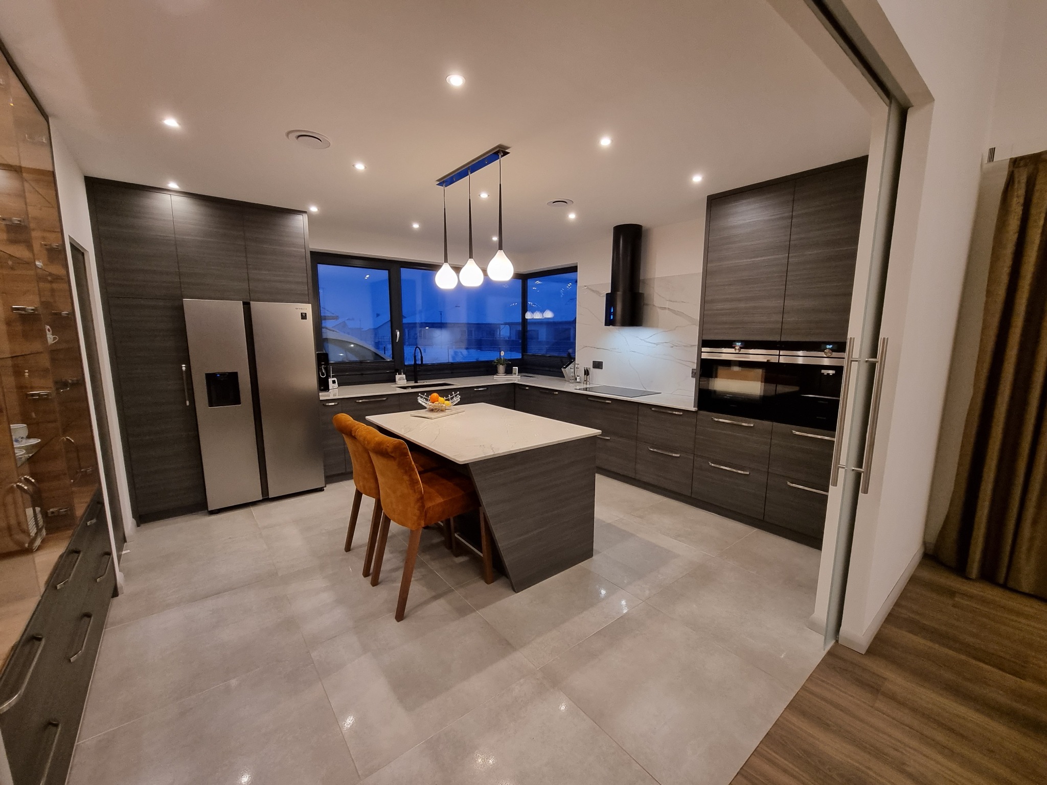 Nowoczesna kuchnia z ciemnymi, drewnopodobnymi szafkami, wyspą z marmurowym blatem i pomarańczowymi krzesłami, oświetlona wiszącymi lampami i punktowymi światłami.