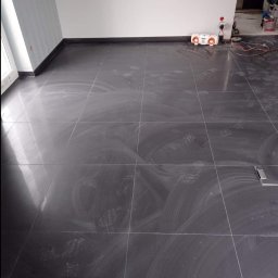 Mar Bud - Świeżo ułożona, błyszcząca, grafitowa podłoga z płytek w pokoju, widoczne ślady po czyszczeniu i odciski butów, białe ściany, czarne listwy przypodłogowe.