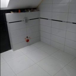 Mar Bud - Wykończona łazienka na poddaszu z białymi, błyszczącymi płytkami i czarnym paskiem dekoracyjnym, widoczny otwór instalacyjny na ścianie.