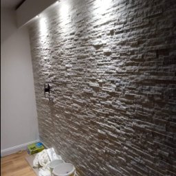 Mar Bud - Ściana wykończona dekoracyjną okładziną z kamienia, oświetlona punktowymi lampami sufitowymi, widoczne akcesoria i narzędzia budowlane w dolnej części kadru.