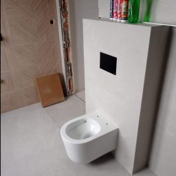 Mar Bud - Świeżo zamontowana podwieszana toaleta z czarnym przyciskiem spłukującym, widoczne silikony i płyn do mycia na obudowie, ściana z dekoracyjną fakturą, płytki podłogowe w odcieniach szarości...