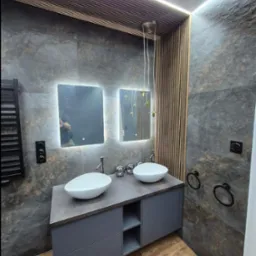 Nowoczesna łazienka z dwoma umywalkami nablatowymi, szarą zabudową, płytkami imitującymi kamień, drewnianymi lamelami na suficie i ścianie, podświetlanymi lustrami oraz drewnopodobną podłogą.