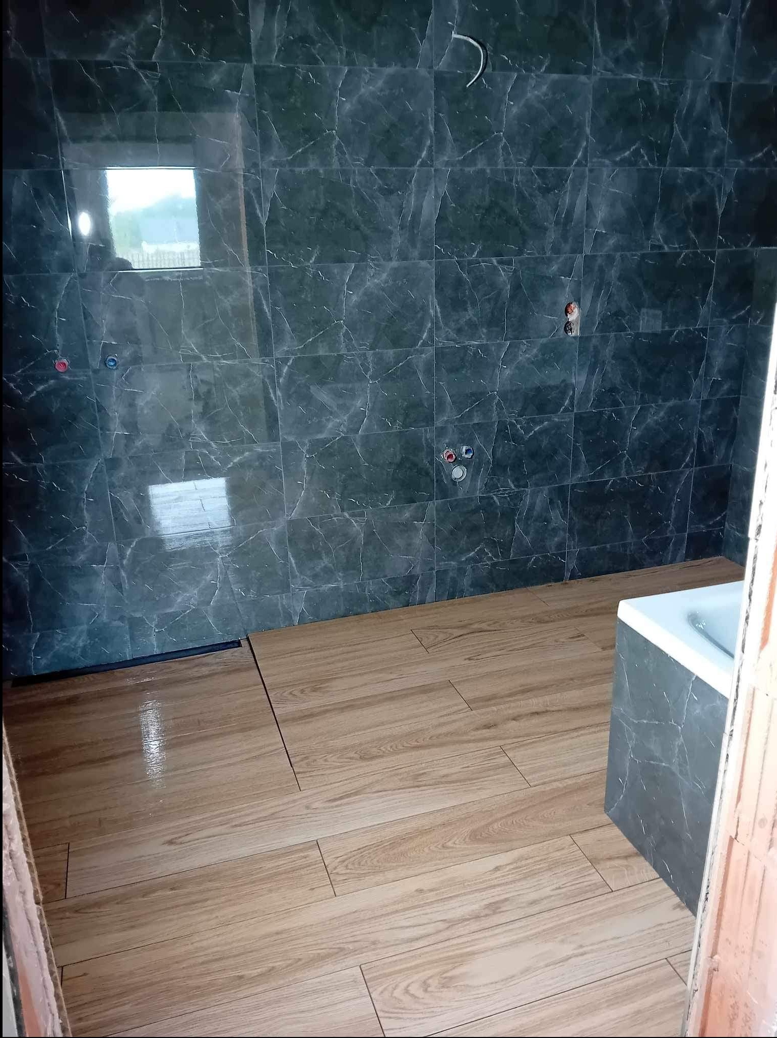 Układanie płytek imitujących drewno na podłodze w łazience z ciemnymi, marmurkowymi płytkami na ścianach. Widoczne przyłącza wodne i fragment wanny.