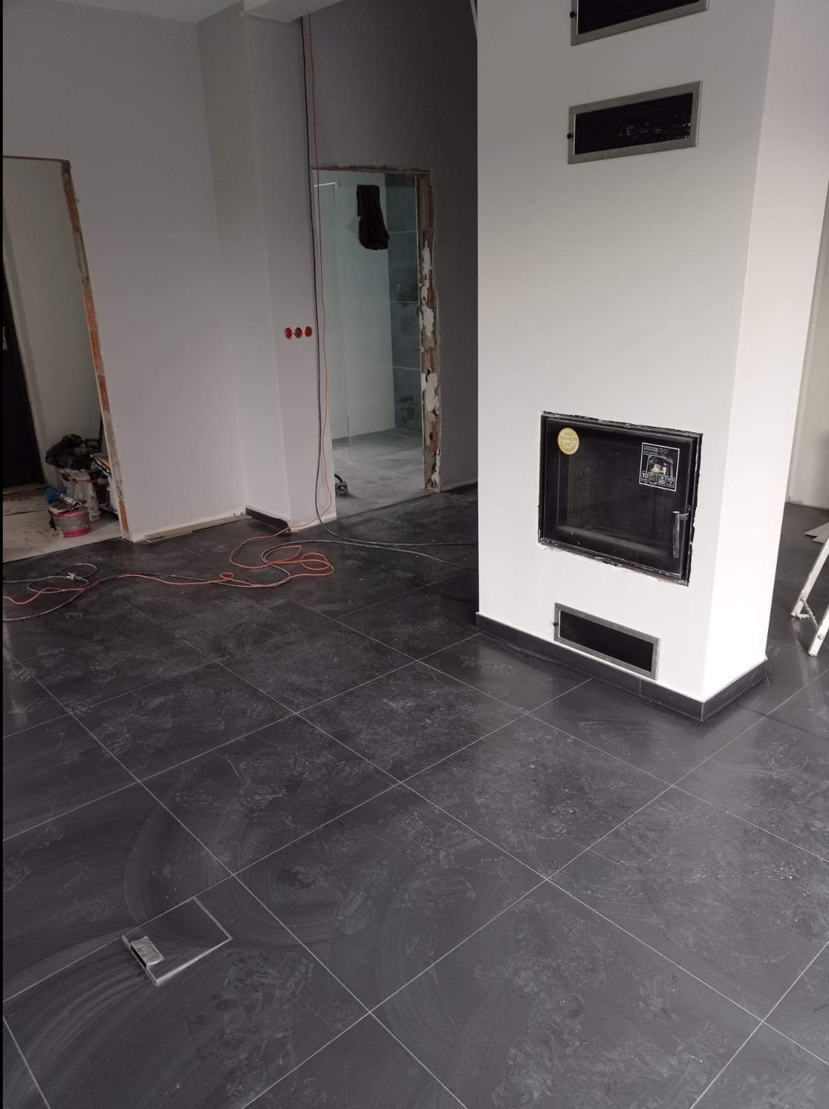 Wykończone podłogi z ciemnej glazury w pokoju z kominkiem, widoczne kable elektryczne i drzwi w trakcie montażu.