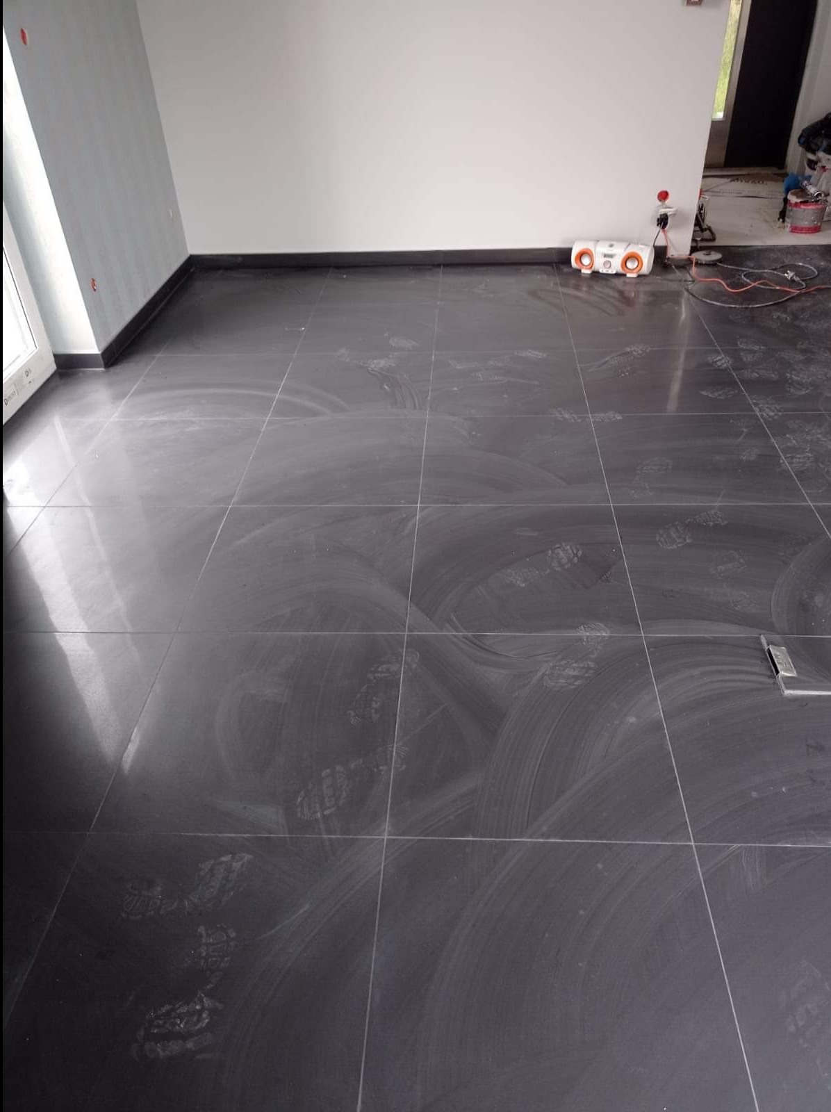 Świeżo ułożona, błyszcząca, grafitowa podłoga z płytek w pokoju, widoczne ślady po czyszczeniu i odciski butów, białe ściany, czarne listwy przypodłogowe.