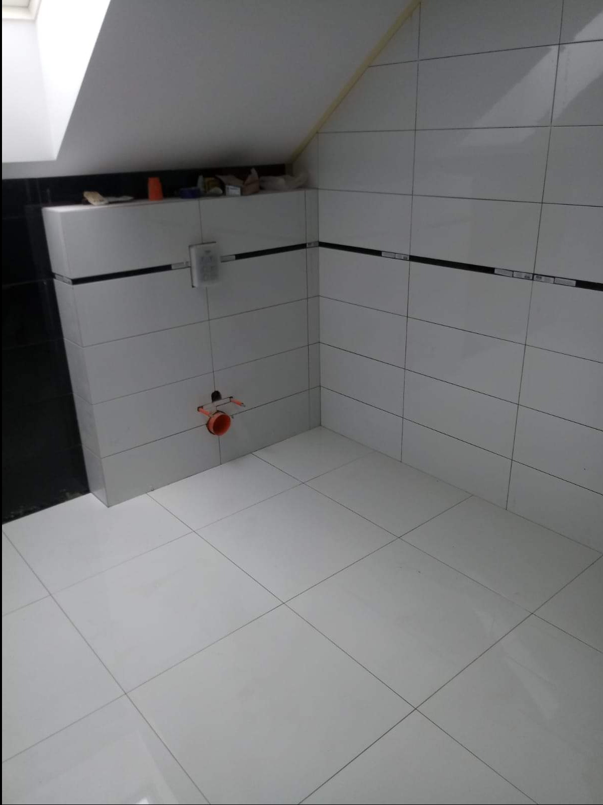 Wykończona łazienka na poddaszu z białymi, błyszczącymi płytkami i czarnym paskiem dekoracyjnym, widoczny otwór instalacyjny na ścianie.