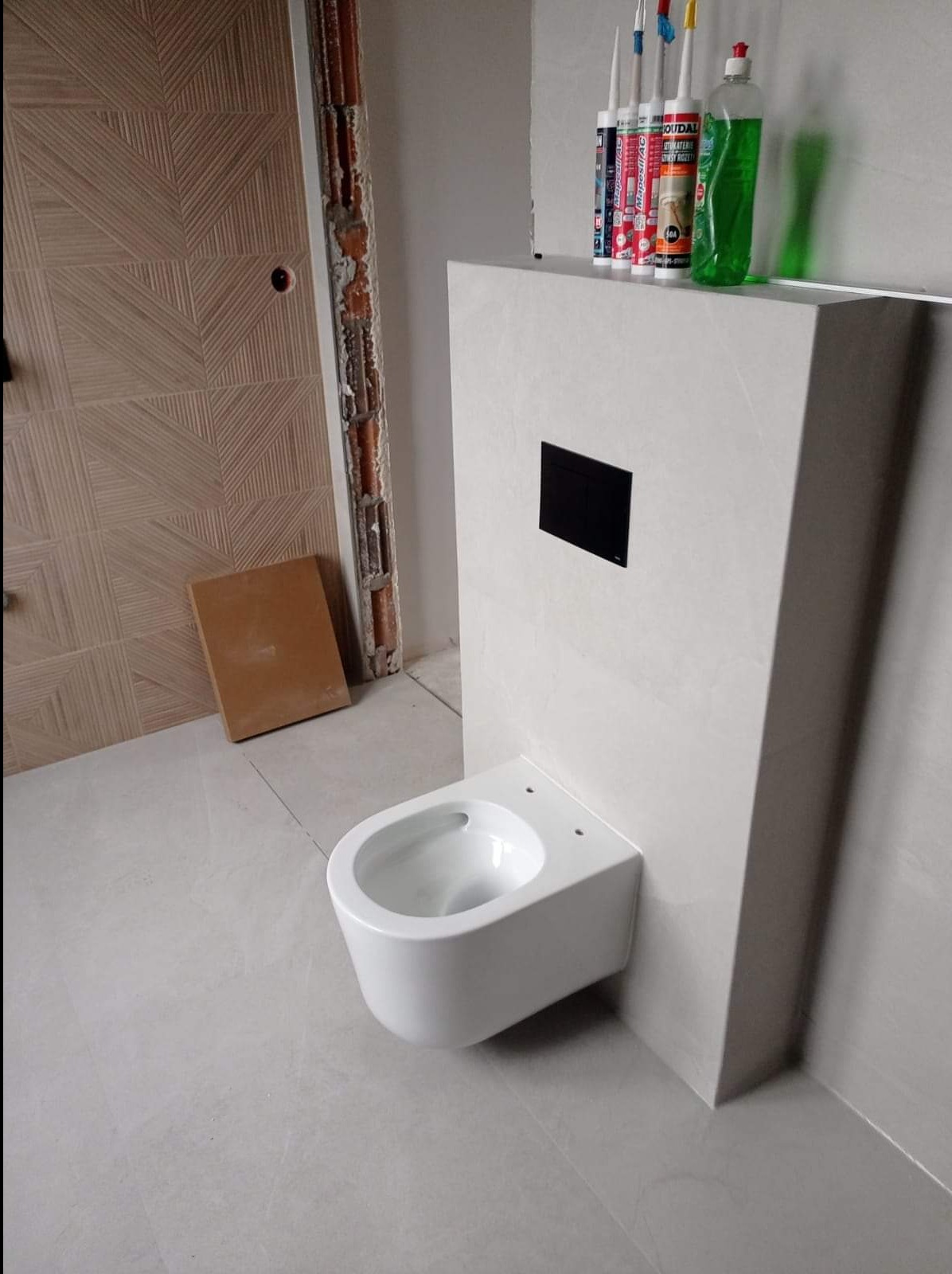 Świeżo zamontowana podwieszana toaleta z czarnym przyciskiem spłukującym, widoczne silikony i płyn do mycia na obudowie, ściana z dekoracyjną fakturą, płytki podłogowe w odcieniach szarości...