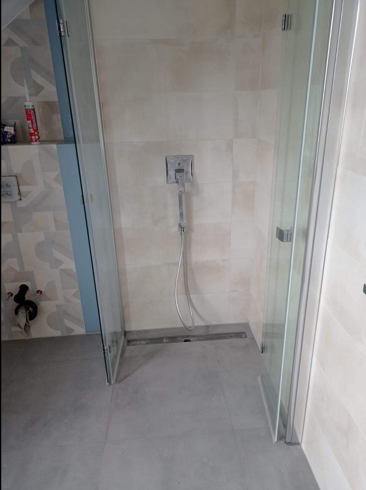 Nowoczesna kabina prysznicowa z szklanymi drzwiami i odpływem liniowym w posadzce, wykończona beżowymi płytkami, z widoczną baterią natryskową i akcesoriami montażowymi.