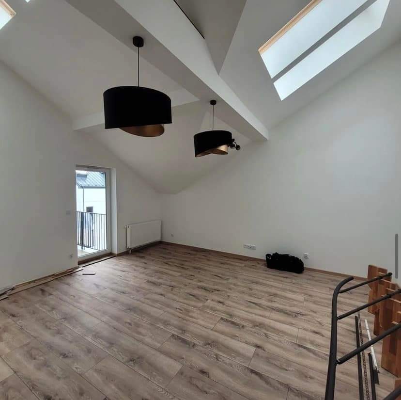 Jasne poddasze po remoncie z drewnianą podłogą, białymi ścianami i czarnymi lampami wiszącymi, widoczne okna dachowe i grzejnik.