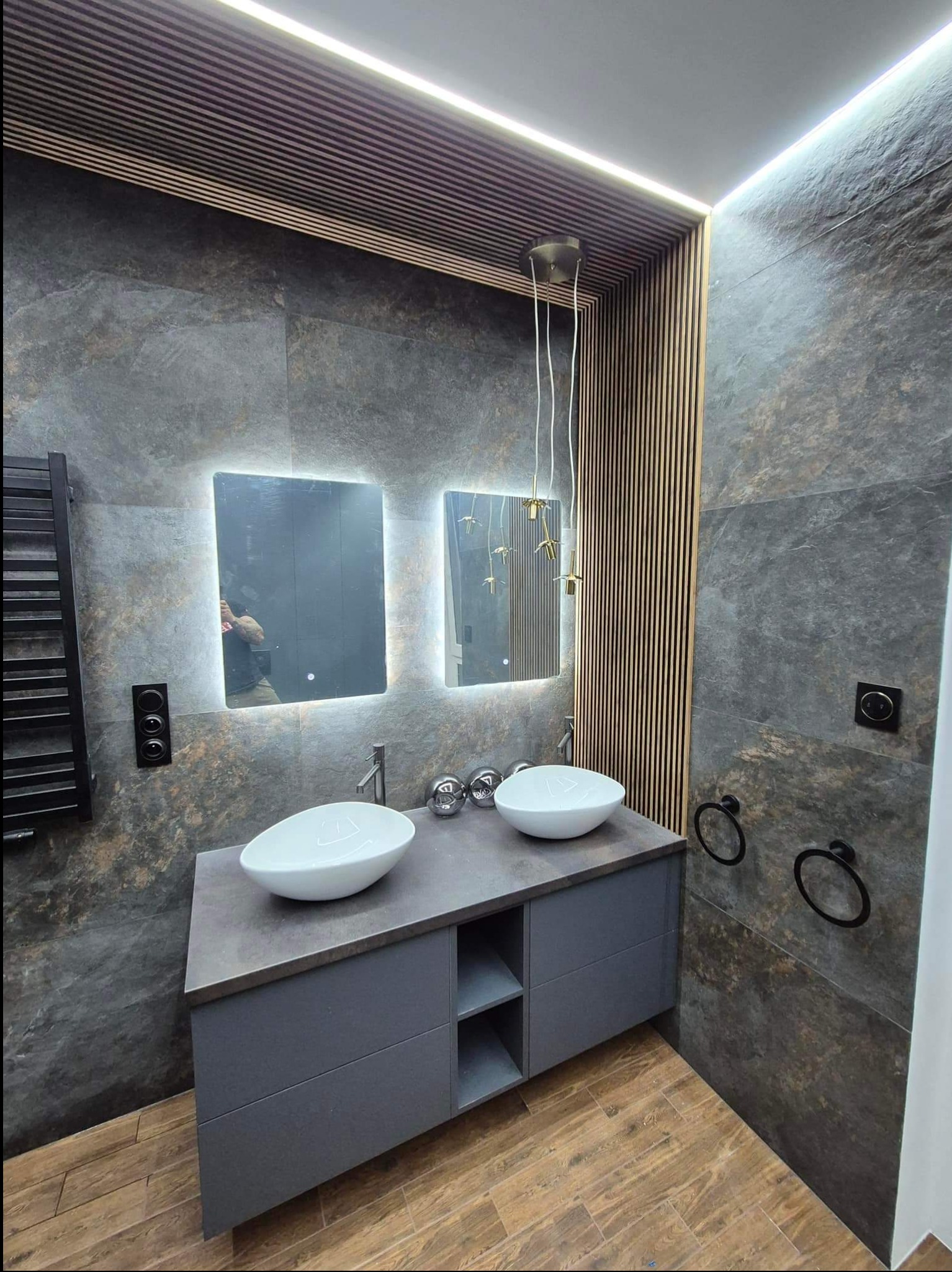 Nowoczesna łazienka z dwoma umywalkami nablatowymi, szarą zabudową, płytkami imitującymi kamień, drewnianymi lamelami na suficie i ścianie, podświetlanymi lustrami oraz drewnopodobną podłogą.