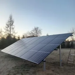 Panel słoneczny zamontowany na metalowej konstrukcji na trawiastym terenie, z widocznymi narzędziami w dolnym rogu i drabiną w tle, sfotografowany o zmierzchu.