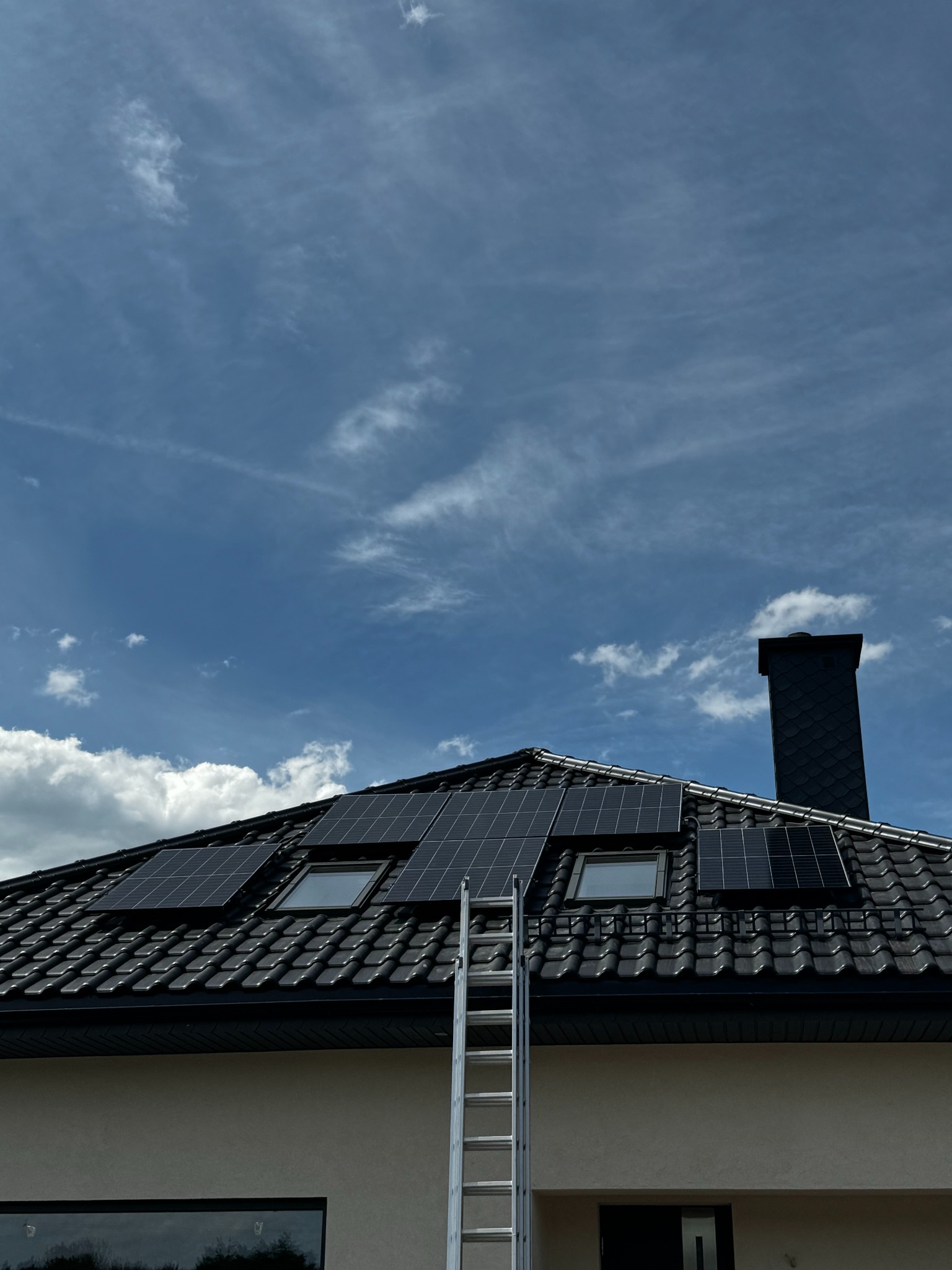 Czarne panele słoneczne zamontowane na dachu z czarnej dachówki, widoczne okna dachowe i komin, drabina oparta o dach, błękitne niebo z chmurami w tle.