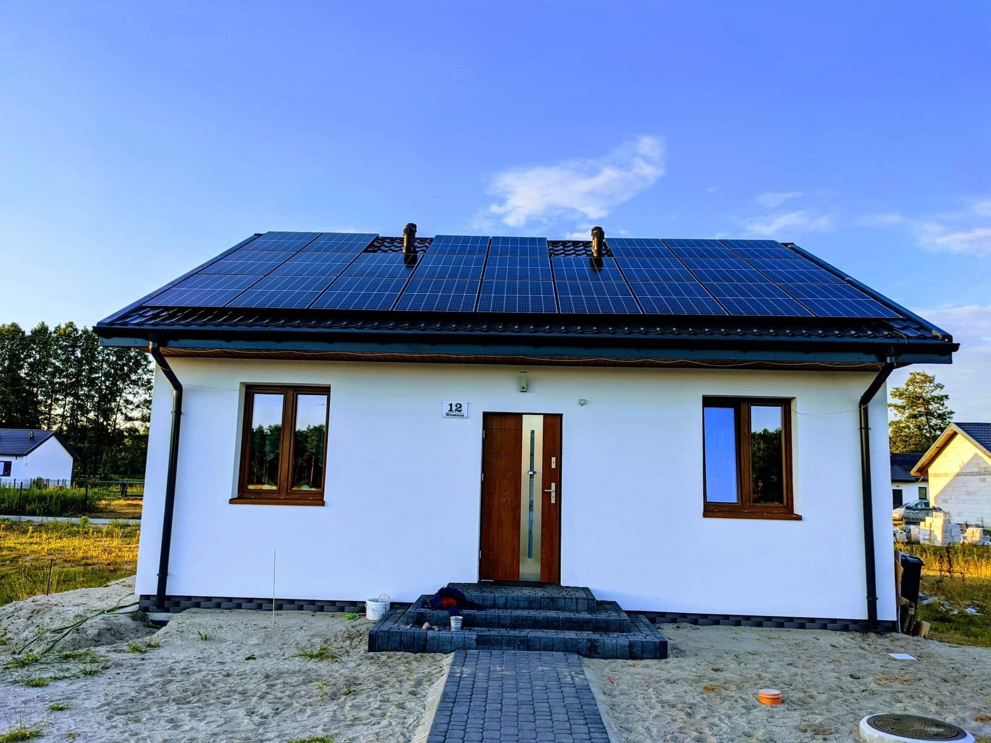 Biały dom jednorodzinny z ciemnymi panelami słonecznymi na dachu, widoczne dwa kominy wentylacyjne, brązowe ramy okienne i częściowo otwarte drzwi wejściowe z numerem '12'.