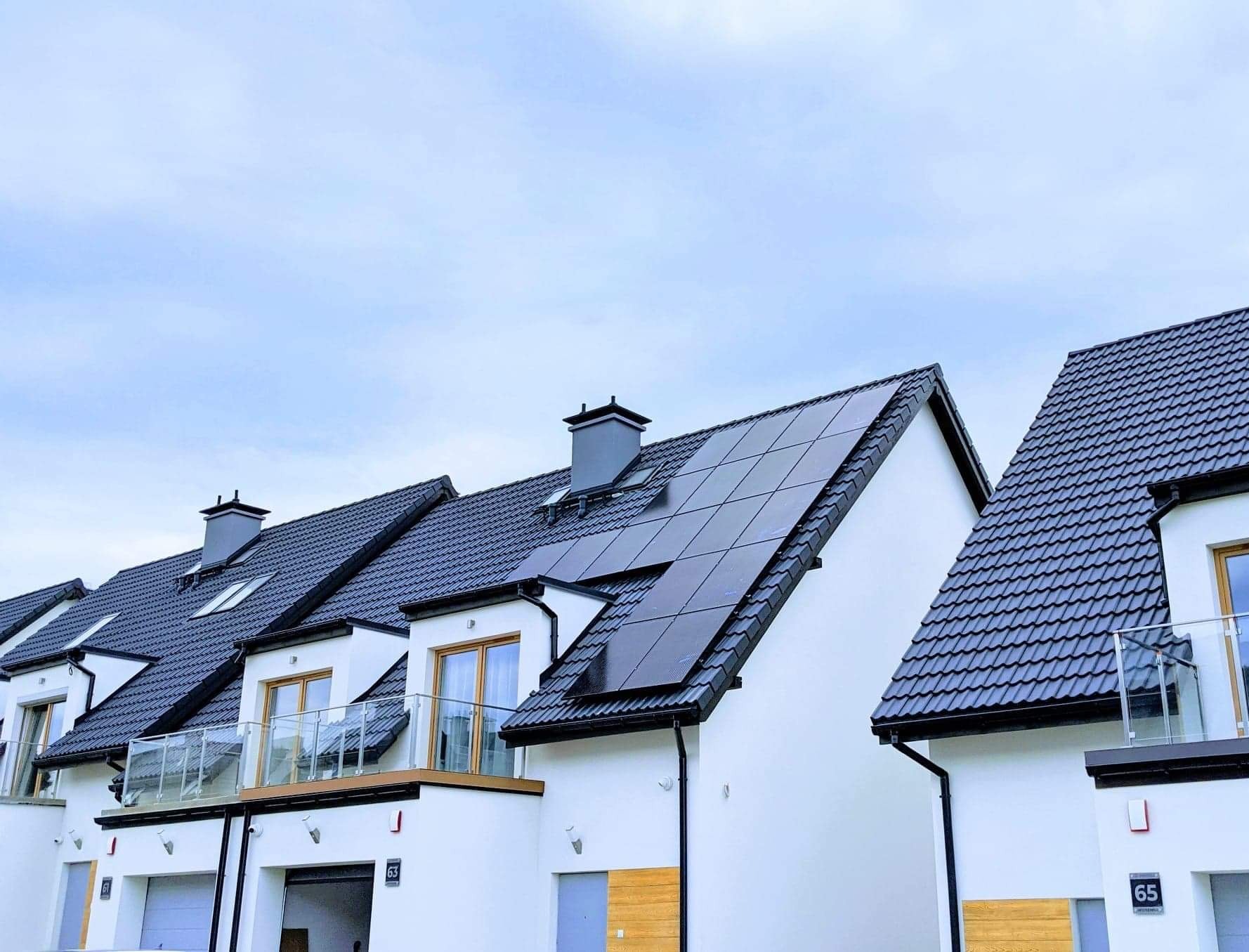 Panele słoneczne zamontowane na dachu szeregowego domu z czarną dachówką, widok z dołu na tle pochmurnego nieba.