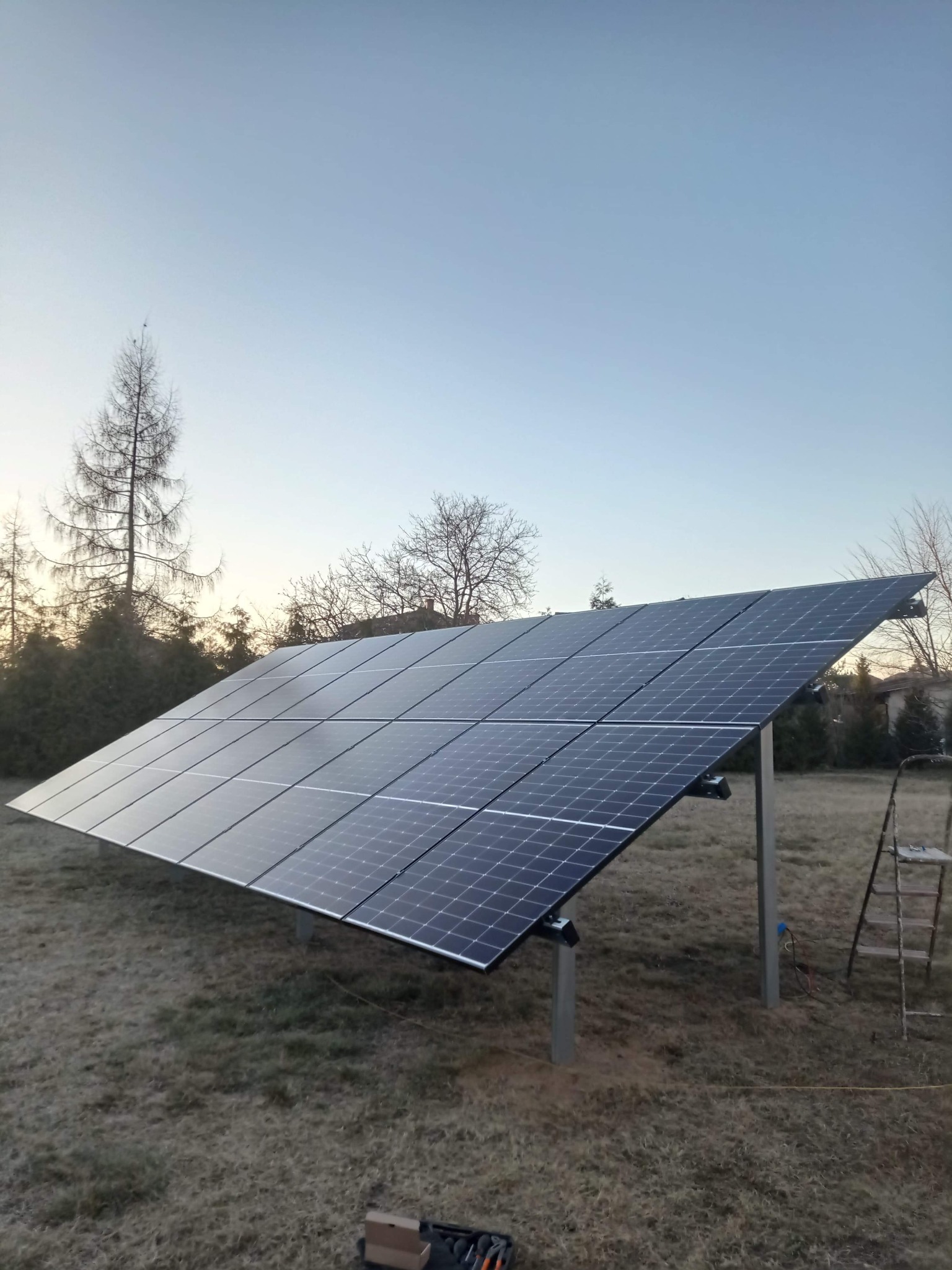 Panel słoneczny zamontowany na metalowej konstrukcji na trawiastym terenie, z widocznymi narzędziami w dolnym rogu i drabiną w tle, sfotografowany o zmierzchu.