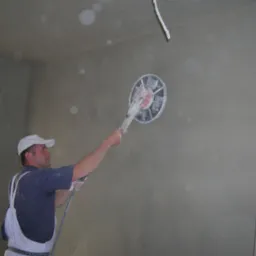 Pracownik w białych spodniach roboczych i czapce wyrównuje świeżo nałożony tynk na ścianie za pomocą elektrycznej szlifierki z okrągłą tarczą, widoczne drobne nierówności na powierzchni ściany...