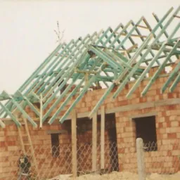 Surowy stan domu w trakcie budowy: ceglana konstrukcja ścian z otworami okiennymi, drewniana konstrukcja dachu z pomalowanymi na niebiesko elementami, widok przez metalowe ogrodzenie.