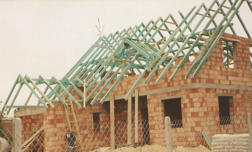 Surowy stan domu w trakcie budowy: ceglana konstrukcja ścian z otworami okiennymi, drewniana konstrukcja dachu z pomalowanymi na niebiesko elementami, widok przez metalowe ogrodzenie.