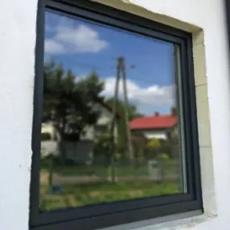 Nowo zamontowane okno z czarną ramą w surowym otworze ściennym, odbijające błękitne niebo i zieleń drzew.