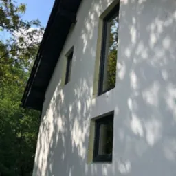 Elewacja domu w Nędzy z trzema oknami o czarnych ramach w różnej wielkości, na tle zieleni i błękitnego nieba, z widocznymi cieniami liści na ścianie.