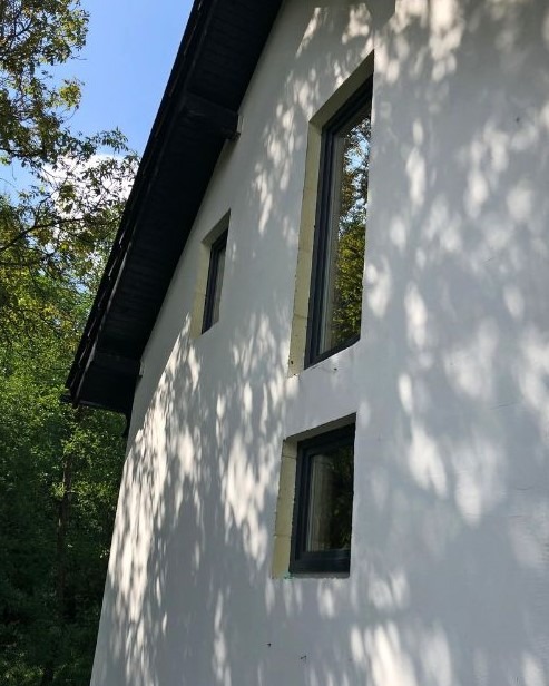 Elewacja domu w Nędzy z trzema oknami o czarnych ramach w różnej wielkości, na tle zieleni i błękitnego nieba, z widocznymi cieniami liści na ścianie.