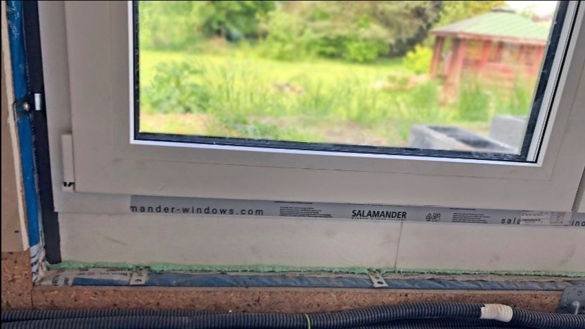 Detal montażu okna z widoczną pianką uszczelniającą, folią ochronną z napisem 'mander-windows.com' oraz fragmentem ogrodu z altaną w tle.