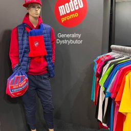 Everglow Paweł Świąć - Manekin w czerwonej bluzie i czapce z daszkiem, granatowej kamizelce i spodniach, z szalikiem i torbą z logo RKS Raków Częstochowa, na tle ściany z logo Modeo Promo i wieszaka z kolorowymi koszulkami.