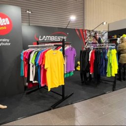 Everglow Paweł Świąć - Stoisko targowe z odzieżą promocyjną: manekin w czerwonym dresie i niebieskiej kamizelce, w tle wieszaki z kolorowymi t-shirtami i osoba oglądająca ubrania.