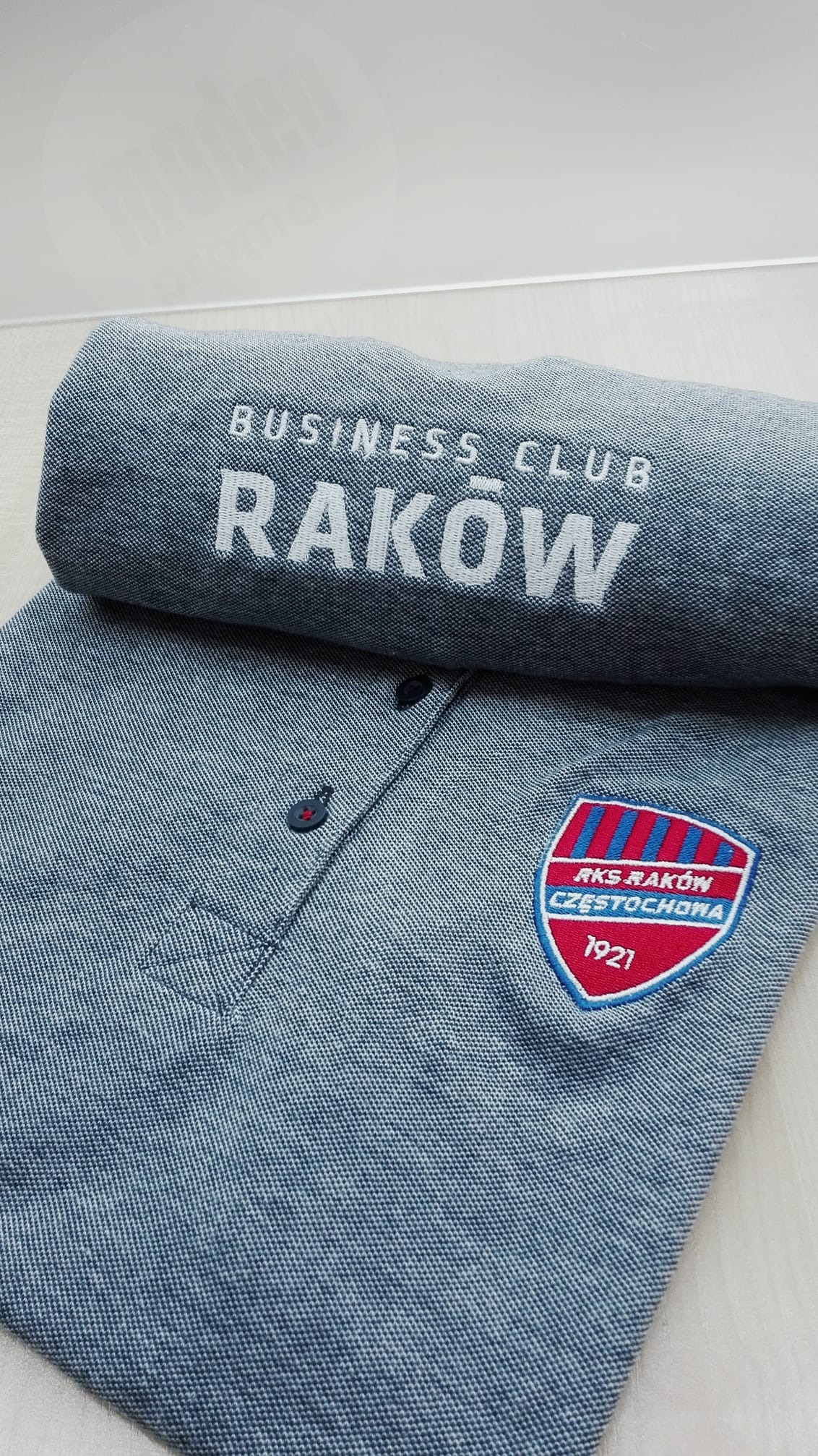 Jasnoniebieska koszula polo z haftowanym herbem RKS Raków Częstochowa i białym napisem 'BUSINESS CLUB RAKÓW' na rękawie