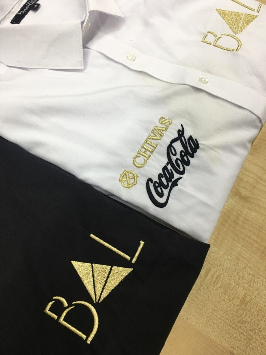 Biała koszula i czarna bluza z wyhaftowanymi złotymi i czarnymi logotypami marek Chivas, Coca-Cola oraz inicjałami 'BVT'.