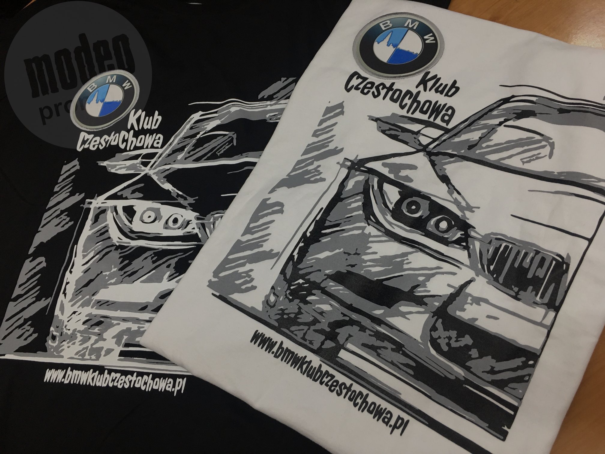 Dwa T-shirty: czarny i biały, z graficznym nadrukiem stylizowanego przodu samochodu BMW i logo klubu BMW Częstochowa, leżące na drewnianej powierzchni.