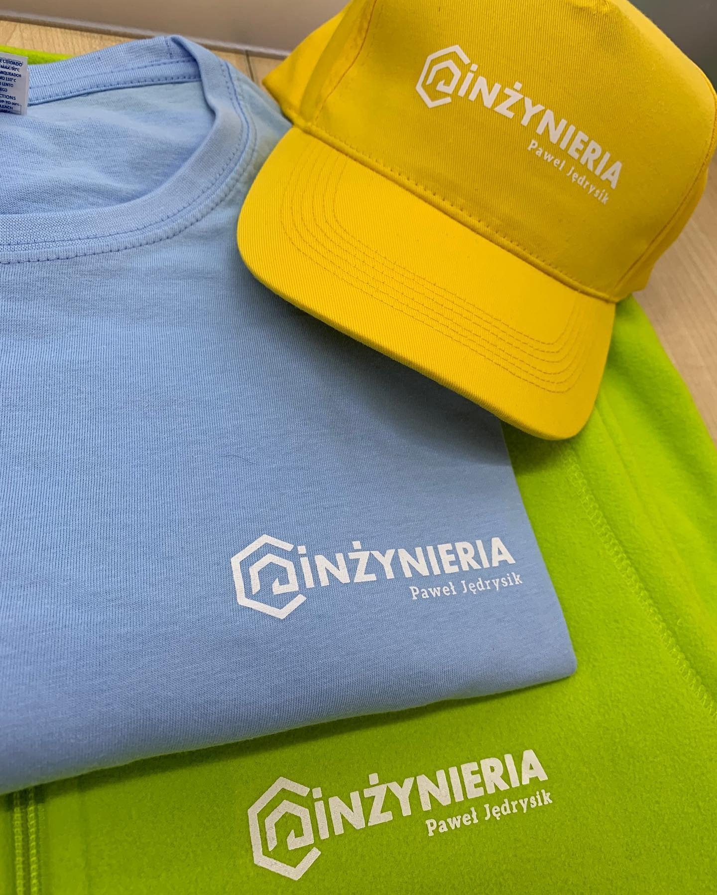 Jasnoniebieska koszulka, limonkowy polar i żółta czapka z daszkiem z białym nadrukiem logo firmy 'Inżynieria Paweł Jędrysik', leżące na drewnianym podłożu.