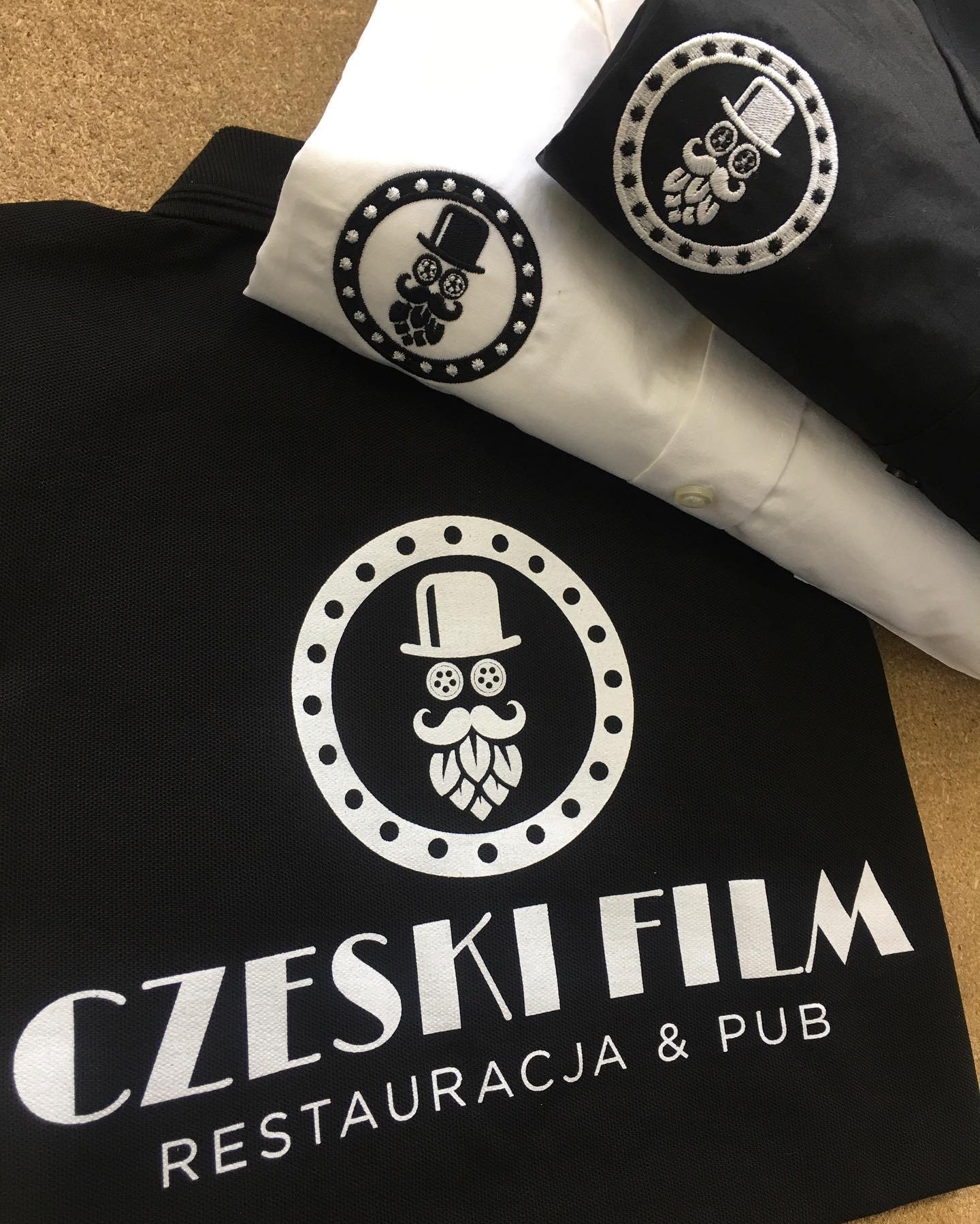 Czarne polo z białym nadrukiem logo restauracji i pubu, obok biała koszula z wyhaftowanym małym logo na piersi.