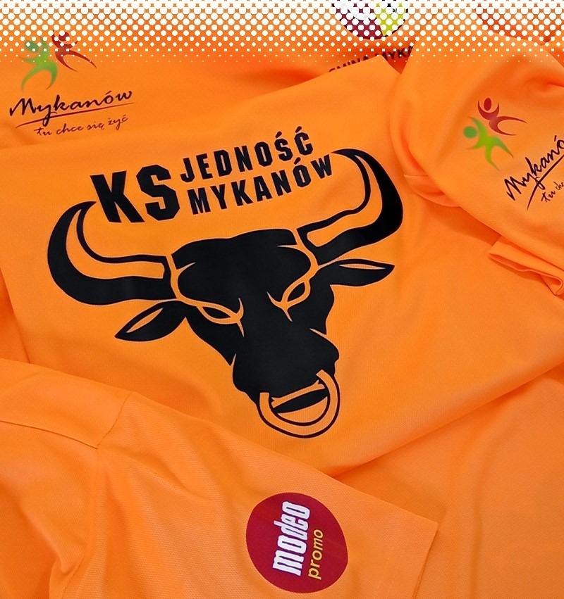 Pomarańczowa odzież sportowa z czarnym nadrukiem głowy byka, napisem 'KS JEDNOŚĆ MYKANÓW' oraz logo 'Mykanów' i okrągłą naklejką 'modeo promo' na rękawie.