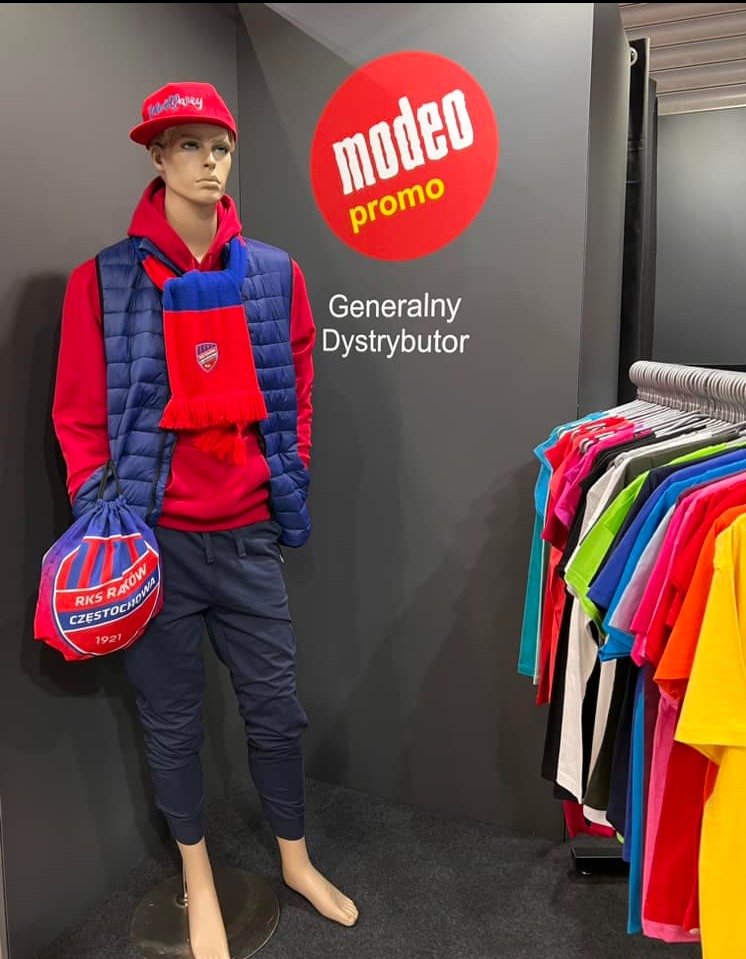 Manekin w czerwonej bluzie i czapce z daszkiem, granatowej kamizelce i spodniach, z szalikiem i torbą z logo RKS Raków Częstochowa, na tle ściany z logo Modeo Promo i wieszaka z kolorowymi koszulkami.