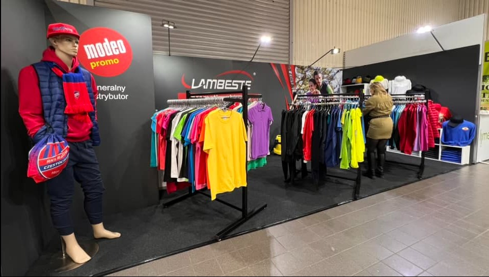 Stoisko targowe z odzieżą promocyjną: manekin w czerwonym dresie i niebieskiej kamizelce, w tle wieszaki z kolorowymi t-shirtami i osoba oglądająca ubrania.