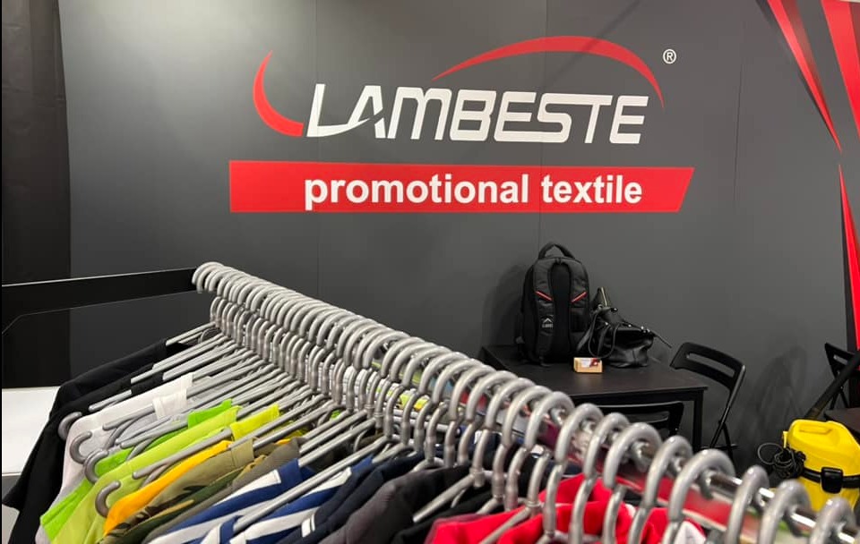 Wieszak z kolorowymi koszulkami promocyjnymi pod logo Lambeste promotional textile na szarym tle, z plecakiem i odkurzaczem w tle.