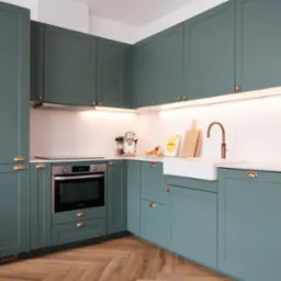 Zielona zabudowa kuchenna z frezowanymi frontami, złotymi uchwytami, białym zlewem farmerskim i blatem z marmuru, oświetlona listwą LED podszafkową.