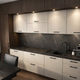NOVESTO - Nowoczesna kuchnia z białymi frontami i ciemnym blatem, uzupełniona o drewnopodobne szafki boczne i sprzęt AGD. Minimalistyczny design z czarnymi uchwytami i marmurowym backsplash.
