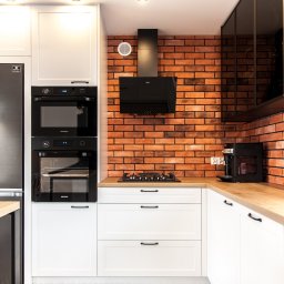 NOVESTO - Nowoczesna kuchnia z białymi szafkami, drewnianym blatem i ścianą z czerwonej cegły. Widoczny sprzęt AGD: lodówka, piekarnik, płyta gazowa i ekspres do kawy.