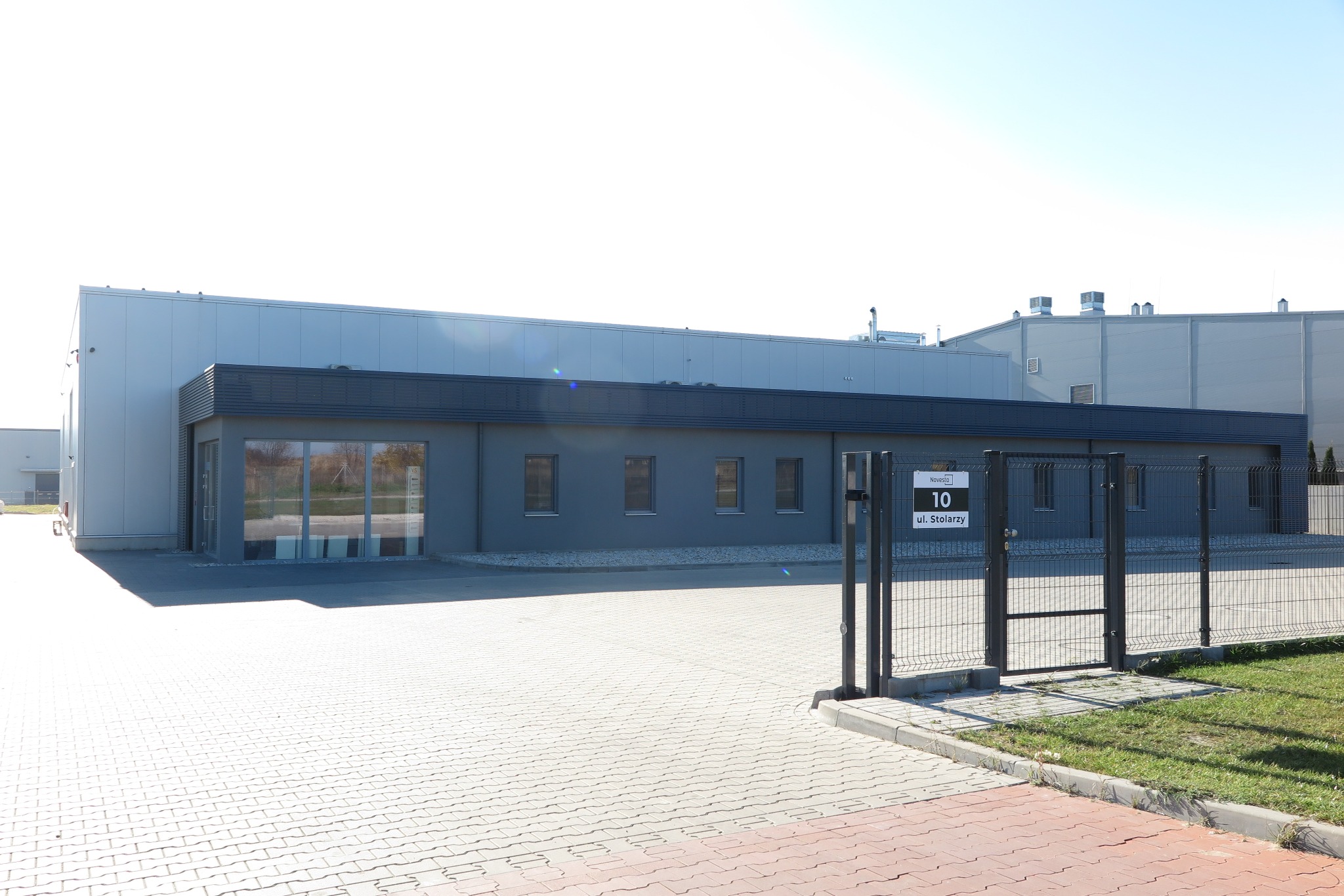 Nowoczesny budynek biurowy w szarym kolorze z dużymi oknami i metalową bramą wejściową z tabliczką adresową. Widok z zewnątrz na plac wyłożony kostką.