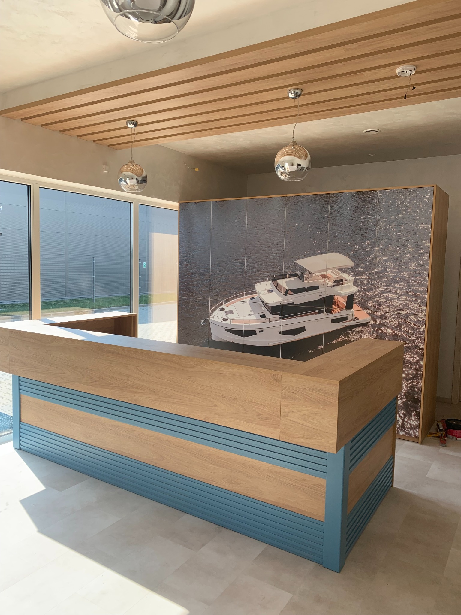 Recepcja z drewnianym blatem i niebieskimi panelami, w tle fototapeta z jachtem, oświetlenie sufitowe z drewnianymi belkami i kulistymi lampami.