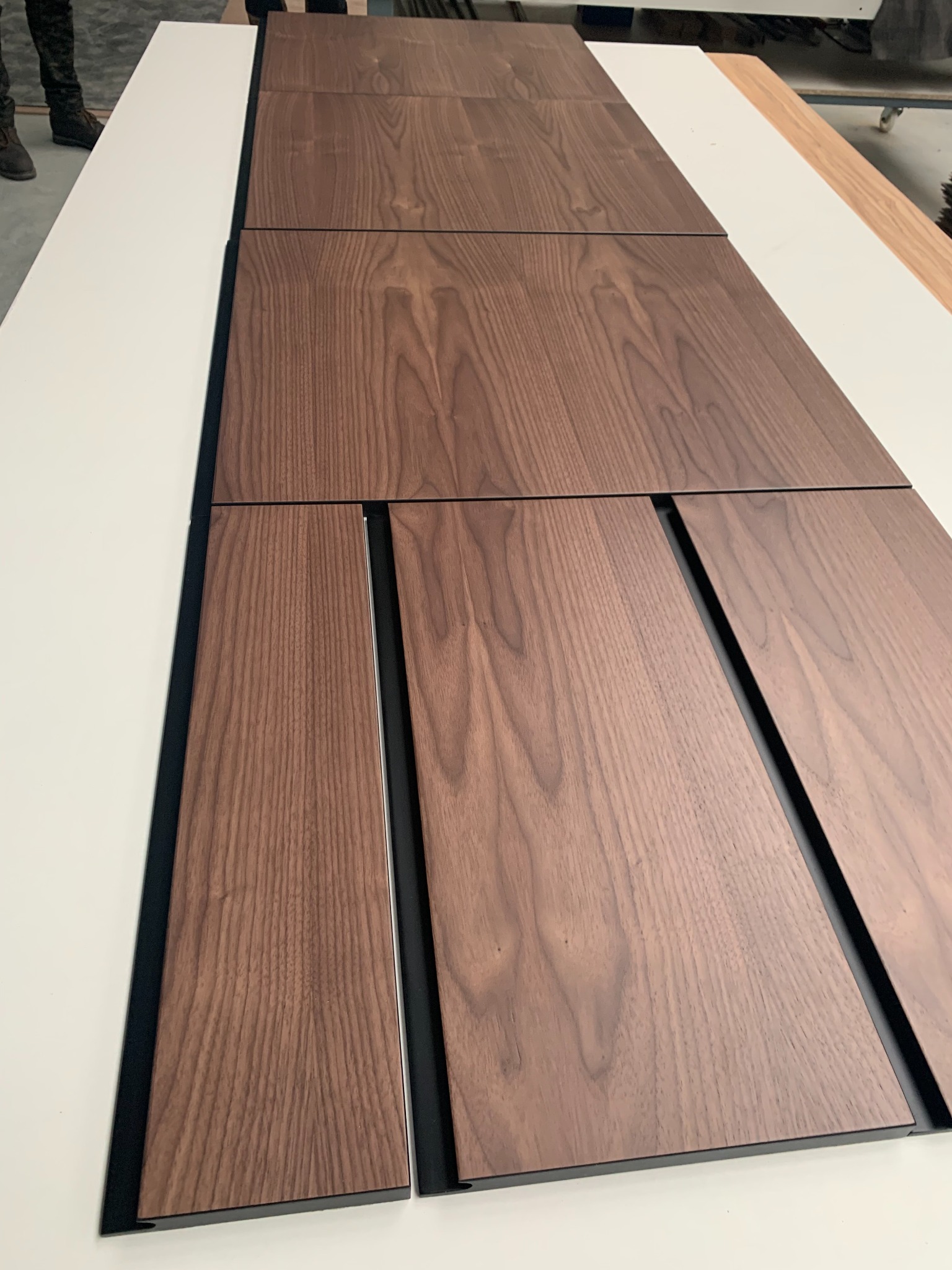 Częściowo złożone fronty szafy z forniru orzechowego z czarnym obramowaniem, leżące na białym blacie roboczym w warsztacie.